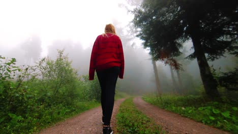 SLOWMO-Low-angle-from-behind,-blonde-girl-in-oversize-sweater-walks-through-a-foggy-forest-trail