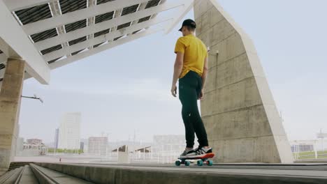 Young-Attractive-Trendy-Man-skateboarding-fast-under-a-solar-panel-on-a-morning-sunny-day-with-an-urban-city-background-in-slow-motion