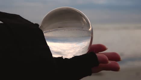 slow-motion-of-a-female-hand-with-fingerless-glove-holding-a-crystal-ball-reflecting-upside-down-a-beach-and-sea-landscape-on-a-cloudy-and-windy-day-of-winter