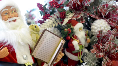 Weihnachtsmann-Spielzeug-Hält-Einen-Stift-Und-Ein-Schönes-Und-Freches-Notizbuch-Mit-Weihnachtsschmuck-Im-Hintergrund