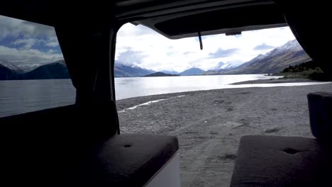 Young-traveler-opening-door-and-showing-exterior-and-interior-of-motorhome-by-beautiful-blue-Lake-Wakatipu,-Queenstown,-New-Zealand-with-mountains-fresh-snow-cloudy-sky-in-background