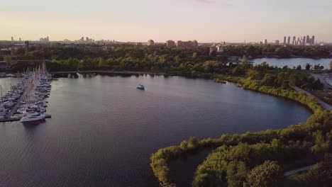 Sonnenuntergang-Aus-Der-Luft,-Absteigende-Aufnahme-Eines-Segelboots-Und-Des-Jachthafen-Yachtclub-Docks-In-Der-Seebucht,-Umgeben-Von-Windigen-Grünen-Bäumen-Mit-Der-Skyline-Der-Stadtgebäude-Im-Hintergrund-In-Toronto,-Ontario,-Kanada