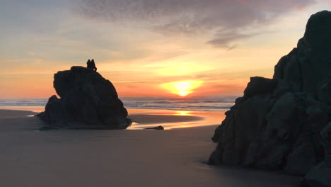 Two-people-sitting-on-a-tall-rock-at-Devil's-Kitchen,-which-is-a-part-of-Bandon-Beach-State-Park-at-the-Oregon-Coast,-enjoying-a-peaceful-and-beautiful-sunset