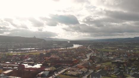Right-rotating-aerial-drone-over-outskirts-of-small-city-with-silhouette-castle-on-hill-in-background-with-high-clouds-and-river