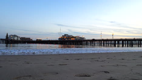 A-scenic-California-sand-beach-with-Stearns-Wharf-pier-beyond-the-ocean-waves-illuminated-by-lanterns-and-lights-at-sunset-in-Santa-Barbara