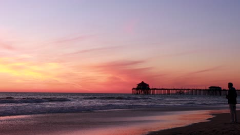 A-man-takes-a-picture-of-the-beach-during-a-gorgeous-yellow,-orange,-pink-and-blue-sunset-with-the-Huntington-Beach-Pier-in-the-background-at-Surf-City-USA-California