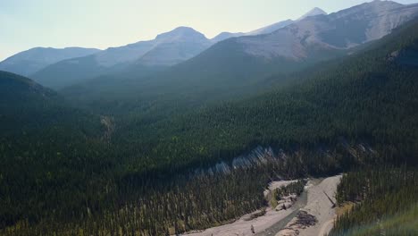 Aerial-Daytime-Wide-Shot-Flying-Forward-Over-A-Summer-Pine-Forest-And-A-Swift-Steep-River-And-Rocky-Mountain-Peaks-in-Alberta-Canada