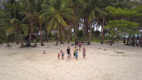 Aerial-static-shot-of-young-caucasian-man-and-filipino-kids-waving-at-drone-from-beautiful-island-with-palm-trees-in-the-Philippines