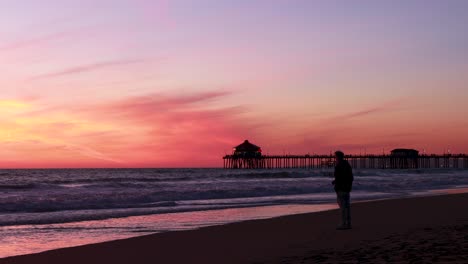 A-man-enjoys-his-vacation-at-the-beach-during-a-gorgeous-red,-purple,-tangerine,-pink-and-blue-sunset-with-the-Huntington-Beach-Pier-in-the-background-at-Surf-City-USA-California
