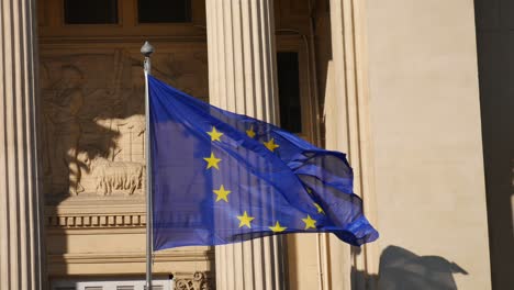 Bandera-Europea-Moviéndose-En-El-Viento-Frente-A-Un-Edificio-Inspirado-En-Las-Antiguas-Columnas-Griegas-Con-Su-Sombra-En-Una-Pared