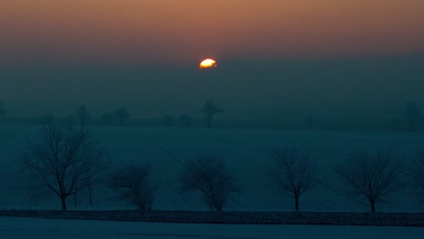 Sunset-time-lapse-on-a-snowy-landscape-with-fields-and-trees,-camera-is-zoomed-and-follows-the-sun