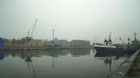 Blue-fisherman's-ship-docked-at-Port-of-Liepaja-in-foggy-day,-dry-cargo-wagons-and-port-crane-in-background,-reflections-in-water,-wide-shot
