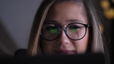 A-face-of-young-woman-shown-from-behind-a-screen,-she-is-scrolling-images-in-browser,-screen-is-reflected-in-her-glasses,-she-smiles-than-is-focused