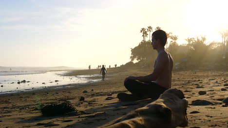 A-handsome-man-deep-breathing-and-meditating-at-sunset-on-a-picturesque-California-beach-with-palm-trees-in-silhouette