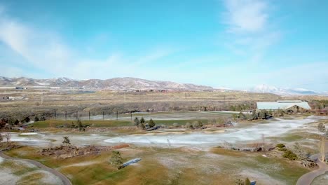 Aerial-view-of-a-golf-course-in-winter-with-a-light-dusting-of-snow-on-the-ground,-mountains-in-the-background-and-wind-turbines