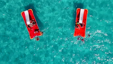 Aerial-shot-of-two-red-pedal-boats-with-tourists-in-crystal-clear-blue-waters-of-a-sandy-beach-at-a-holiday-resort