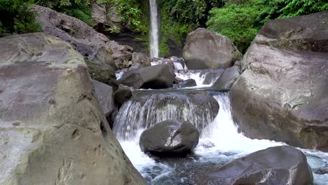 Close-up-of-river-water-rushing-over-boulders-and-rocks-with-a-tropical-waterfall-in-the-background-in-a-rainforest-in-Asia
