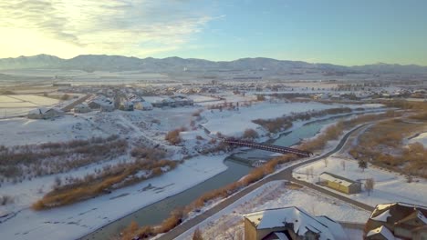 Aerial-view-of-a-neighborhood-with-children-sledding-down-a-slope-near-a-river-and-bridge-with-mountains-and-a-sunset-in-the-background