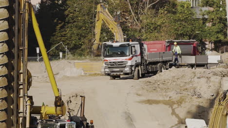 Excavadora-Descargando-Tierra-En-Un-Camión-En-Un-Sitio-De-Construcción-En-Viena,-Austria-En-Cámara-Lenta-Con-Un-Trabajador-Pasando