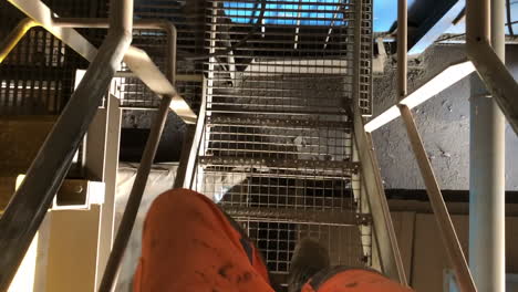 Shot-of-a-worker-his-feet-while-he-walks-down-the-stairs-in-a-factory,-wearing-high-visible-orange-safety-clothing