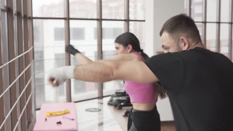 Young-athletic-woman-in-a-fistical-gloves-is-training-boxing-in-a-fitness-room-with-her-male-couch