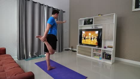 A-man-is-stretching-his-legs-while-standing-in-front-of-a-television-in-his-living-room-following-a-fintess-programme-trying-to-burn-some-calories