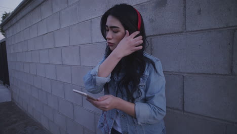 Young-woman-looking-down-her-phone-in-the-alleyway,-next-to-wall,-wearing-red-headphones---medium-shot