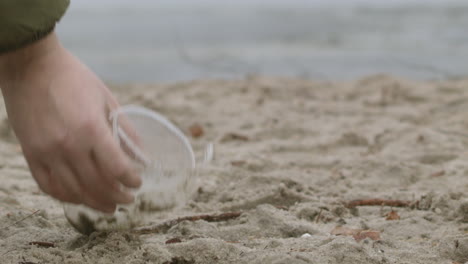 Die-Hand-Eines-Mannes-Hebt-Einen-Leeren-Plastikbecher-Am-Strand