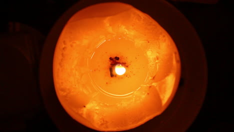 An-orange-and-red-candle-with-a-litted-shaky-flame-and-melted-wax-on-dark-background-viewed-closely-from-above
