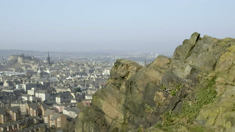 Pan-right-to-left-from-male-and-female-hikers-on-the-top-of-Salisbury-Crags-over-to-Edinburgh-city-centre-below-with-Edinburgh-castle-in-the-background-on-a-sunny-day,-Scotland