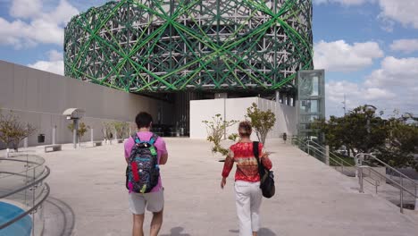 Panning-to-the-right-as-two-people-walk-past-the-camera-toward-the-Great-Museum-of-the-Mayan-World-in-Merida,-Yucatan,-Mexico