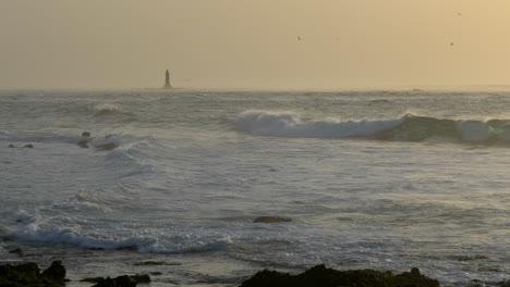 Goldene-Meereslandschaft-Bei-Sonnenuntergang,-Entfernter-Leuchtturm,-Wellen-In-Zeitlupe,-Die-Am-Strand-Krachen