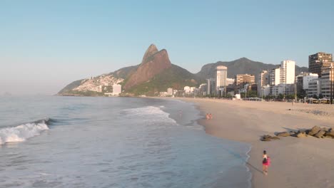 Sideways-aerial-movement-accompanying-a-wave-coming-in-on-coastal-city-beach-of-Rio-de-Janeiro-during-early-morning-golden-hour-seen-from-above-the-ocean