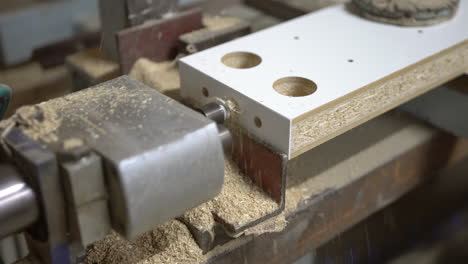 Horizontal-technical-drill-bit-goes-in-and-out-into-chipboard-wooden-element,-producing-a-lot-of-flying-sawdust-arround