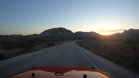 Driving-toward-the-sunrise-in-a-red-vehicle-in-the-desert-with-mountains-visible-in-silhouette