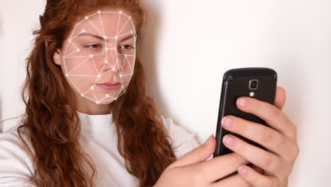 Close-up-of-a-young-woman-with-red-hair,-with-a-white-shirt-on-a-white-background,-while-holding-a-phone-in-front-of-her-face-with-facial-recognition-high-tech-animation-with-a-glow-effect-on-her-face