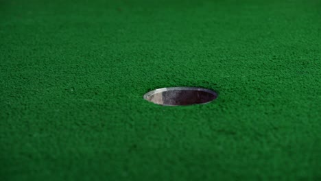 An-orange-mini-golf-ball-exits-a-pipe-in-a-wall-and-rolls-over-for-a-hole-in-one-shot
