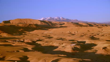 Toma-Panorámica-Del-Paisaje-Seco-Del-Desierto-Con-Montañas-Cubiertas-De-Nieve-En-El-Fondo-En-Marruecos