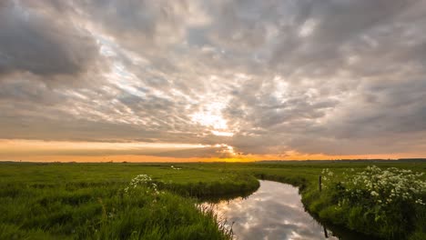 Dramatic-movement-of-clouds-over-a-green-field-and-river-at-sunset
