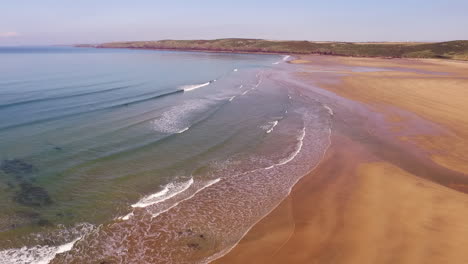 Aerial-shot-looking-across-the-sandy-beach-at-Freshwater-west-in-Wales,-with-small-waves-and-sunny-skies,-while-panning-right