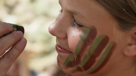 Extreme-Close-Up-of-Smiling-Girl-Gets-Her-Face-Painted-With-Green-And-Brown-Colors-War-Paint-By-Another-Person