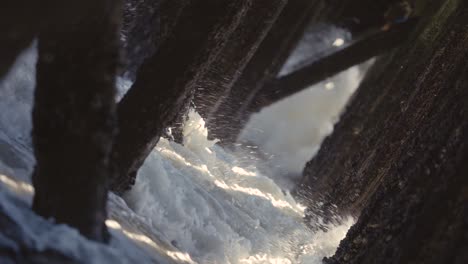 Waves-crashing-under-a-pier,-old-wooden-pillings-with-sunbeams-coming-through
