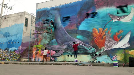 Graffiti-Artists-Outside-Spray-Painting-A-Large-Wall-With-A-Seascape-With-A-Whale-In-An-Urban-Setting-In-Mexico-During-The-Day