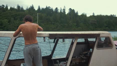 Topless-young-male-walks-into-frame-scrapes-old-mastic-from-timber-boat-cabin-roof-planking