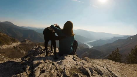Girl-sitting-and-cudling-black-labrador-dog-on-a-mountain-with-beautiful-lake-canyon-in-the-background