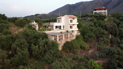 Aerial-Pull-Away-from-Luxury-Greek-Villa-with-Pool-with-Mountains-in-Background-and-Green-Foliage-in-Foreground---Car-on-Drive