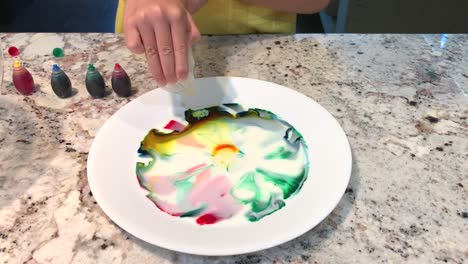 Small-hands-dropping-liquid-detergent-onto-the-plate-causing-a-cool-chemical-reaction-that-mixes-up-and-spreads-out-the-colors-into-a-beautiful-pattern-of-swirls