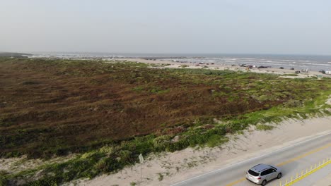 Aerial-decent-from-view-of-the-beach-and-grasslands,-landing-in-parking-lot-with-2-vehicles-passing