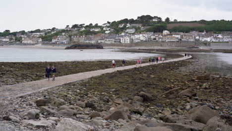 Tourists-walking-on-the-causeway-that-leads-to-Marazion-from-the-english-medioeval-castle-and-church-of-St-Michael's-Mount-in-Cornwall-on-a-cloudy-spring-day,-4k-footage