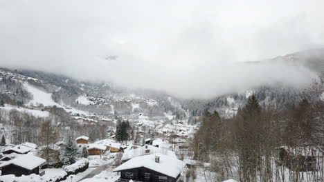 Drone-shot-flying-through-snowy-ski-village-with-low-cloud,-mountains-trees,-and-chalets-in-the-background
